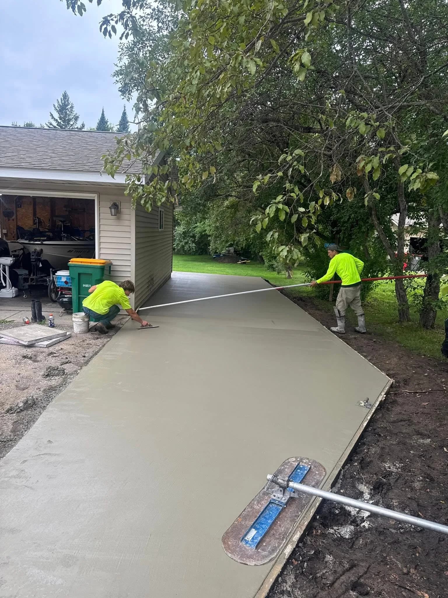 Two concrete workers in high-visibility shirts pouring a residential driveway. One edges the perimeter while the other uses a bull float to level the wet concrete.